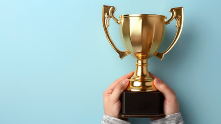 Close-up image of hands holding a golden trophy against a light blue background.の素材