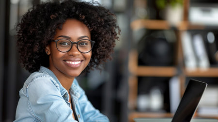 A young woman wearing glasses smiles warmly as she works on a laptop in a modern office setting.の素材