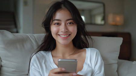 A woman in a white shirt sits on a couch, smiling and looking at the camera while using her smartphone.の素材
