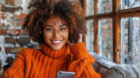 A young woman in an orange sweater smiles while looking at her phone.の素材
