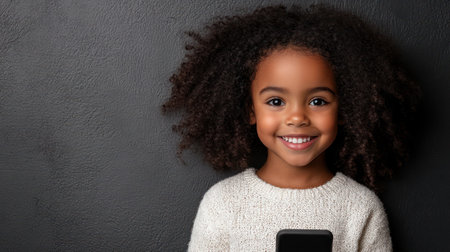 A young girl with curly hair is happily holding a smartphone, smiling at the camera in a warm indoor environment.の素材