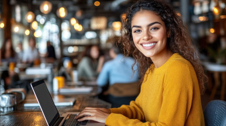 A cheerful young woman engages with her laptop in a bustling cafe, surrounded by fellow customers.の素材
