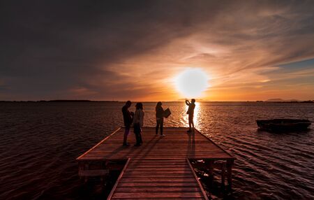Four young guys and girls take a picture of the sunset on the water with a smart phone, from a wood pier in the coast of the Stagnone, a low sea water next to Marsala, province of Trapani, Sicilyの写真素材