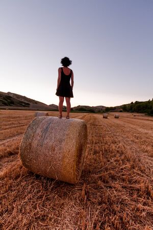 Caucasian 30s brunette woman, standing on a hay bale during Golden hourの写真素材