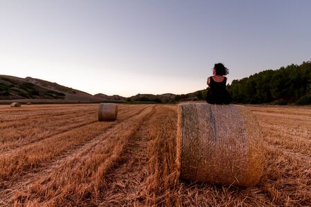 Caucasian 30s brunette woman, sitting on a hay bale during Golden hourの写真素材
