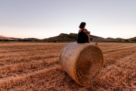 Caucasian 30s brunette woman, sitting on a hay bale during Golden hour