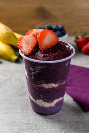 Brazilian frozen aÃ§ai berry ice cream on a plastic cup with strawberry and oatmeal flakes. bananas and strowberries background. wooden desk. Summer menu front view.の写真素材