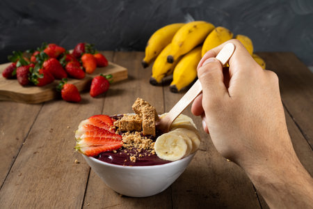Spoon and a White Bowl of Brazilian Frozen AÃ§ai Berry With PaÃ§oca, Strawberry and Banana. on a wooden desk.の写真素材