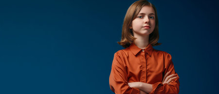 Serious young woman in orange shirt standing with crossed arms on blue backgroundの素材