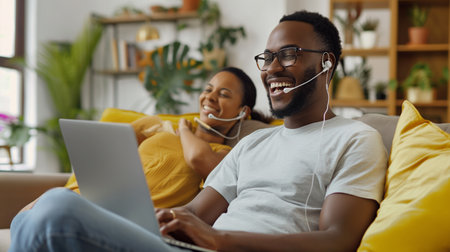 Cheerful african american man in earphones using laptop while sitting on couch at homeの素材