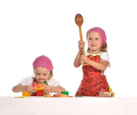 two pretty little twins sisters wearing spotted pink headscarfs and colourful aprons pretending housewifes cooking isolated on white backgroundの写真素材