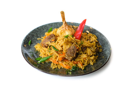 dish of cooked pilaff with rice and meat on plate with head of garlic, pepper and greens on white...