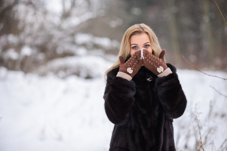 A beautiful smiling blonde girl on the background of a winter snowy forest covers her face with warm brown glovesの写真素材