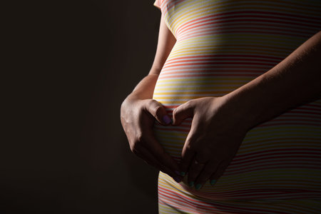 the girl hugs her pregnant belly, folding her hands like a heart. She is in a dress on a dark studio...