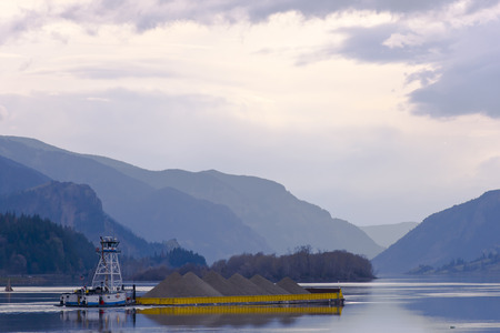 Tugboat pushing a loaded yellow barge on the river surface of the evening against mountains and cloudy skyの写真素材