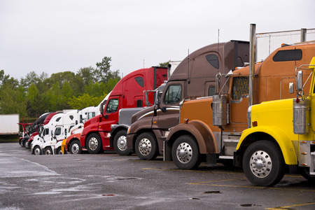 Cabins of multicolored semi trucks lined up on the truck stop after a rain with puddles on the pavement on the background of green trees and gray rainy overcast sky