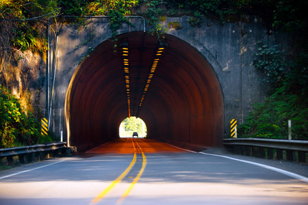 A road with a dividing strip for the opposing traffic flows pass through a long semi-circular concrete tunnel carved in the rockの写真素材