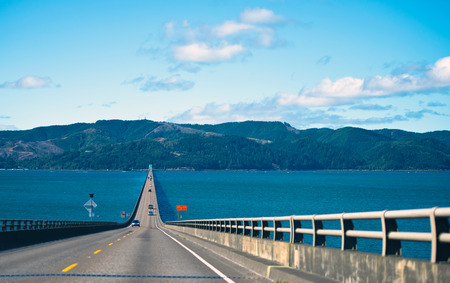 Long spectacular road bridge with several lanes with raised sections and fencing in wide mouth of the Columbia River in Astoria, Pacific, rests on the horizon on the opposite bank of the hilly with trees.の写真素材