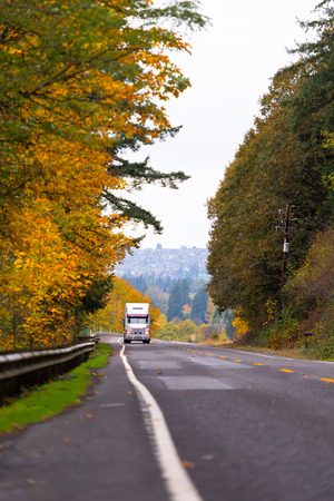 Large big rig classic white semi truck on scenic highway with two lanes with a metal security fence, road markings and yellowed autumn trees on both sides of the road. Against the background of rising above the houses.の写真素材