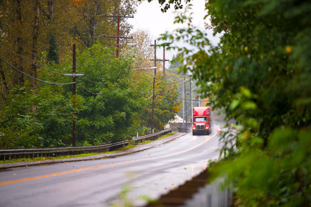Shiny big classic heavy-duty modern red semi truck with a trailer in the rain with wooden poles of...