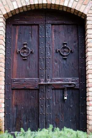 Ancient castle door, dark stained wood with rivets and forged round rings handles and powerful deadbolt closed with a modern lock in the arched wall of the old building, built of brick.のeditorial素材