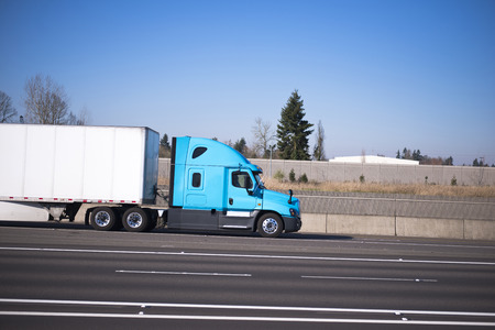 Bright blue modern road train of the semi truck and the dry van trailer streamlined aerodynamic skirt on the highway with lots of traffic lanes separated by markings and concrete fence. Side view.の写真素材