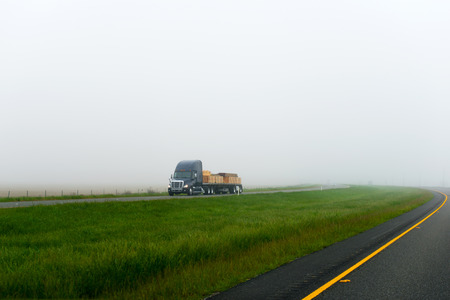 The modern model of a large dark-colored semi truck with a flat bed trailer laden piles of lumber on Interstate highway with a wide dual carriageway with grass moves in a dense fog that covers the other cars and the horizonの写真素材