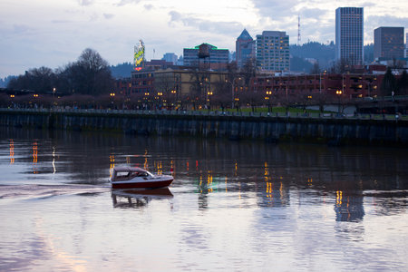 A small boat with a convertible cabin on Willamette River in the heart of Portland with an evening promenade enabled streetlights and office buildings towering above the waterfront walkways reflected lights.のeditorial素材