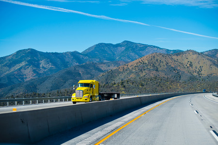 Bright yellow powerful big rig semi truck with a flat bed trailer on the interstate mountain pass highway turn with the mountain tops and blue sky.の写真素材