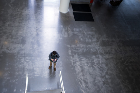 guy with a backpack and a smartphone in hand and headphones standing in the spacious lobby of a modern building with a concrete polished floor with the reflection of the light falling on the floor behind the figure of a young man holding modern technologyの写真素材