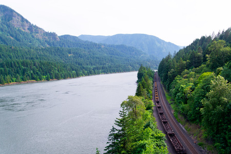 The railway and moving freight train on it was laid along the scenic Columbia River shore is surrounded by greenery of the trees on the slopes of the steep rock formations. Evergreen trees and the waters of the river create a unique beauty of Columbia Gorの写真素材