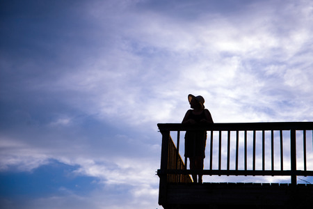 Silhouette of a woman in a hat with a beautiful figure leaned against the balcony の写真素材
