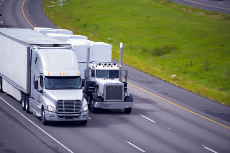 Two heavy big rigs semi trucks of various types and models with different trailers and various cargoes rapidly moving close together on the international highway.