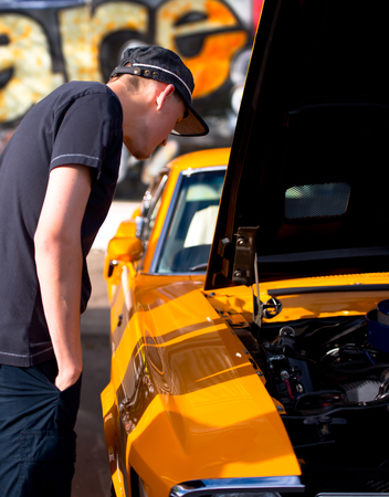 A young man in a cap with hands in pockets examines classic vintage muscle bright yellow car with open hood standing in the street on the day of parade of retro cars.の写真素材