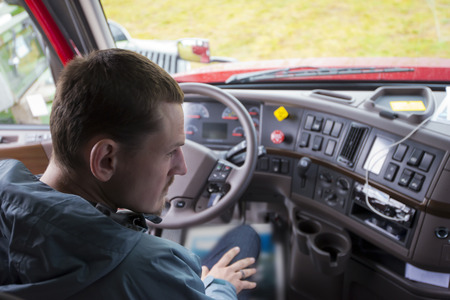 The truck driver sitting in the cab of modern comfort and ergonomic semi truck behind the wheel and interior dashboard with numerous control buttons and switches. The driver of semi truck assessment the situation, looking in the right rearview mirror.の写真素材