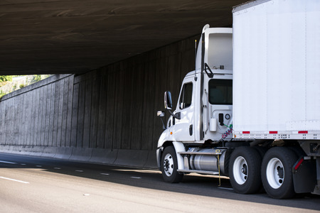 White powerful professional big rig semi truck for local regional transportation transports cargo with dry van trailer on a highway under the concrete bridge with shadow on the side wallの写真素材