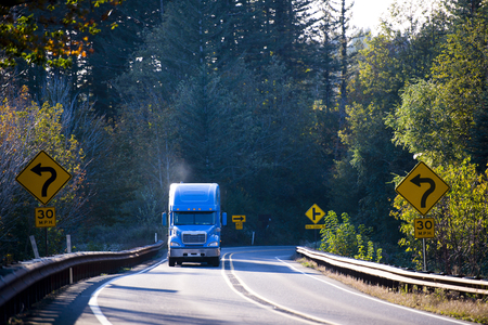 Large American professional powerful truck with smoke from the exhaust pipe, transports goods on the winding road through the mountains, surrounded by yellowing autumn trees illuminated by bright sunshine, with road signs on both sides of the road.の写真素材