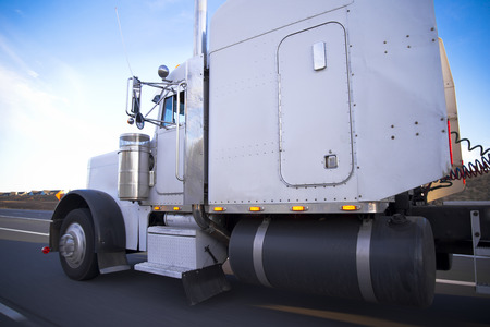 A classic huge, powerful white truck for long distance haulage with a long cab and a huge fuel tank is speeding to its destination on the straight highwayの写真素材