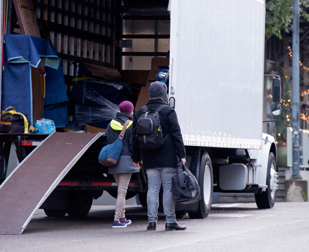 A young couple is transporting goods in a white truck with a boxing trailer and a wheelbarrow ramp to a new place of residence in urban city. The girl and the guy keep the bags in their hands and inspect the furniture to be unloaded.の写真素材
