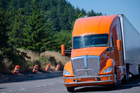Big rig contemporary orange semi truck with chrome grille protection guard and dry van trailer running on green highway with trees on roadside and road working equipment conesの写真素材