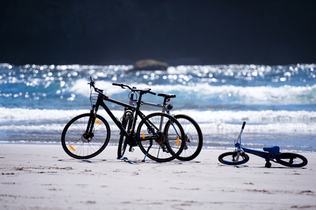Bicycles of the mother of father and child are on the shore of the Pacific Ocean, the waves of which shine and shimmer in the sun, waiting for an exciting fun cycling tripの写真素材