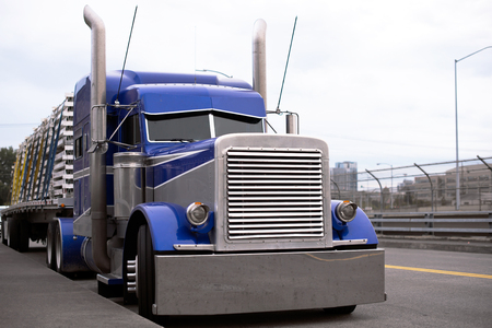 Face of blue American classic popular powerful big rig semi truck tractor with huge grille and chrome accessories and tall exhaust pipes with flat bed trailer going on the road for delivery commercial cargo to destination warehouseの写真素材