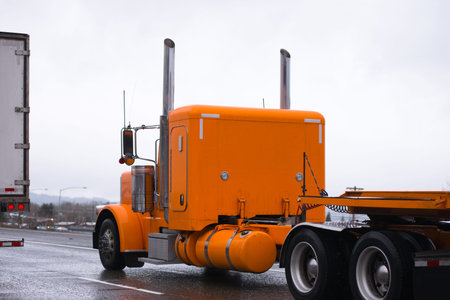 Orange comfort professional American bonnet big rig classic old style semi truck driving with flat bed semi trailer on highway in rain weather for loading cargo for deliveryの写真素材