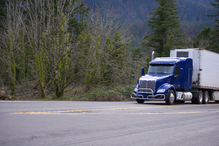 Dark blue American bonnet day cab big rig semi truck with grille guard for local deliveries carry refrigeration semi truck transporting goods on wide road with forest trees backgroundの写真素材