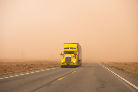 Yellow big rig bonnet powerful semi truck with reefer semi trailer caring cooled cargo going rides during huge strong sandstorm with poor visibility on the road in Utahの写真素材