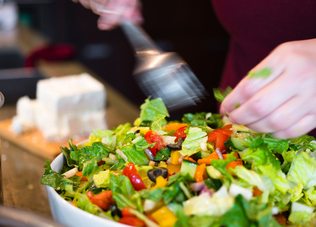 Cook adds diced cheese in the fresh organic healthy low-calories dietary Greek salad with greens salad leaves, red tomatoes, pepper, paprika, cabbage, cucumbers seasoned with olive oil in large bowlの写真素材