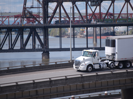 Big rig white day cab semi truck for local delivery transporting cargo in refrigerated semi trailer with refrigerator unit on in on the overpass road along the river with bridge on backgroundの写真素材