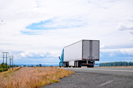 Blue modern bonnet powerful long haul big rig semi truck with dry van semi trailer transport commercial cargo for timely delivery on scenic road with grass forest mountain and cloudy skyの写真素材