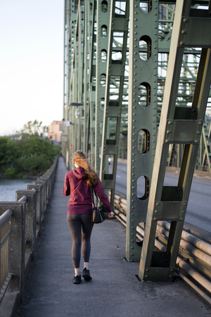 Girl with long hair and bag over her shoulder walks across the Columbia Interstate lift bridge with triangle connected trusses to other side of the river, focusing on communicating on a mobile phoneの写真素材