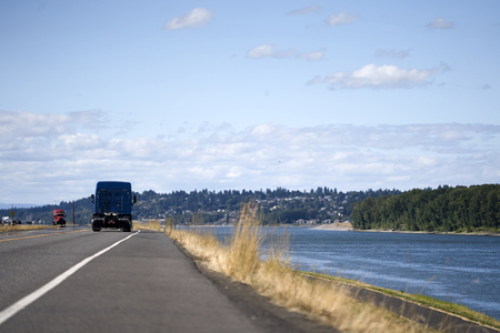 Big rig dark semi truck tractor without of semi trailer going on the road with another traffic along the river to pick up the trailer with cargo and delivery it to destination warehouse on the timeの写真素材